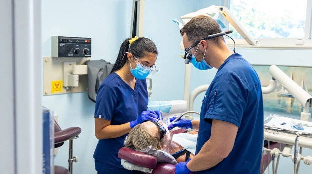 Two dental professionals in blue scrubs examine a patient in a modern dental operatory.